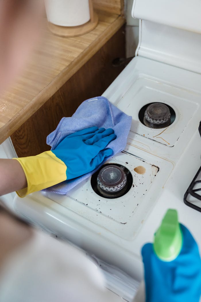 contact-img A person wearing rubber gloves cleaning a dirty gas stove with a cloth and spray bottle.